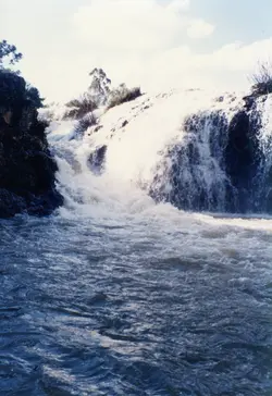 Esta imagem colorida retrata uma cachoeira com grande volume de água caindo sobre rochas e vegetação, formando um rio ou lago na parte inferior. O céu azul com algumas nuvens brancas é visível ao fundo. A qualidade da fotografia e as cores sugerem que pode ser uma imagem do final do século XX, possivelmente de uma área natural em uma região como Videira ou uma pequena cidade brasileira.