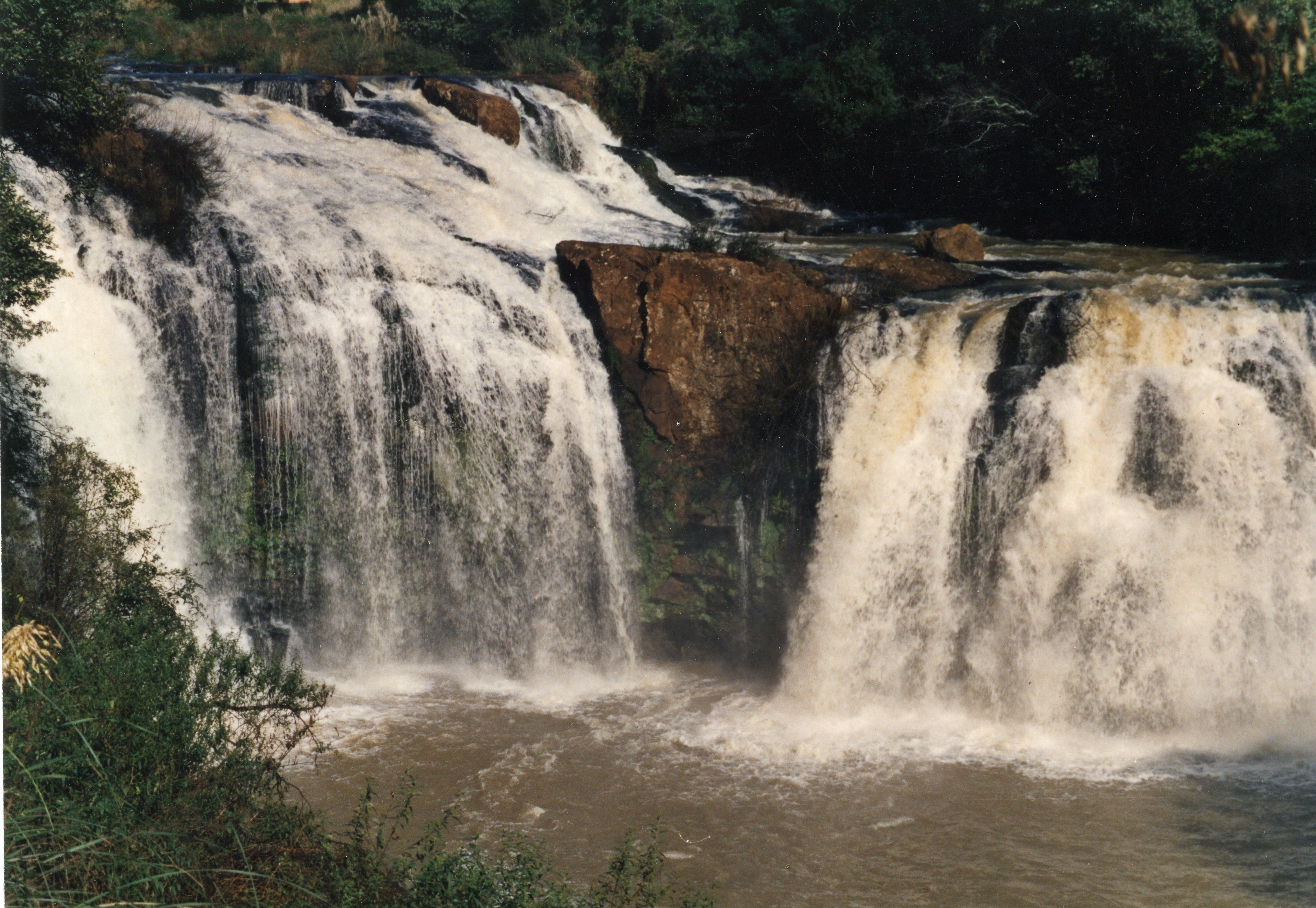 Uma fotografia colorida de uma cachoeira com queda d'água volumosa e rochas expostas, cercada por vegetação densa. A imagem captura a força da natureza em um ambiente possivelmente rural, comum na região de Videira, Santa Catarina.