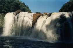Uma fotografia de uma cachoeira larga com queda d'água volumosa cercada por vegetação densa e rochas. A imagem captura a força da natureza em um ambiente rural, possivelmente em uma região de serra brasileira, com uma estética que remete a fotografias de natureza do final do século XX.