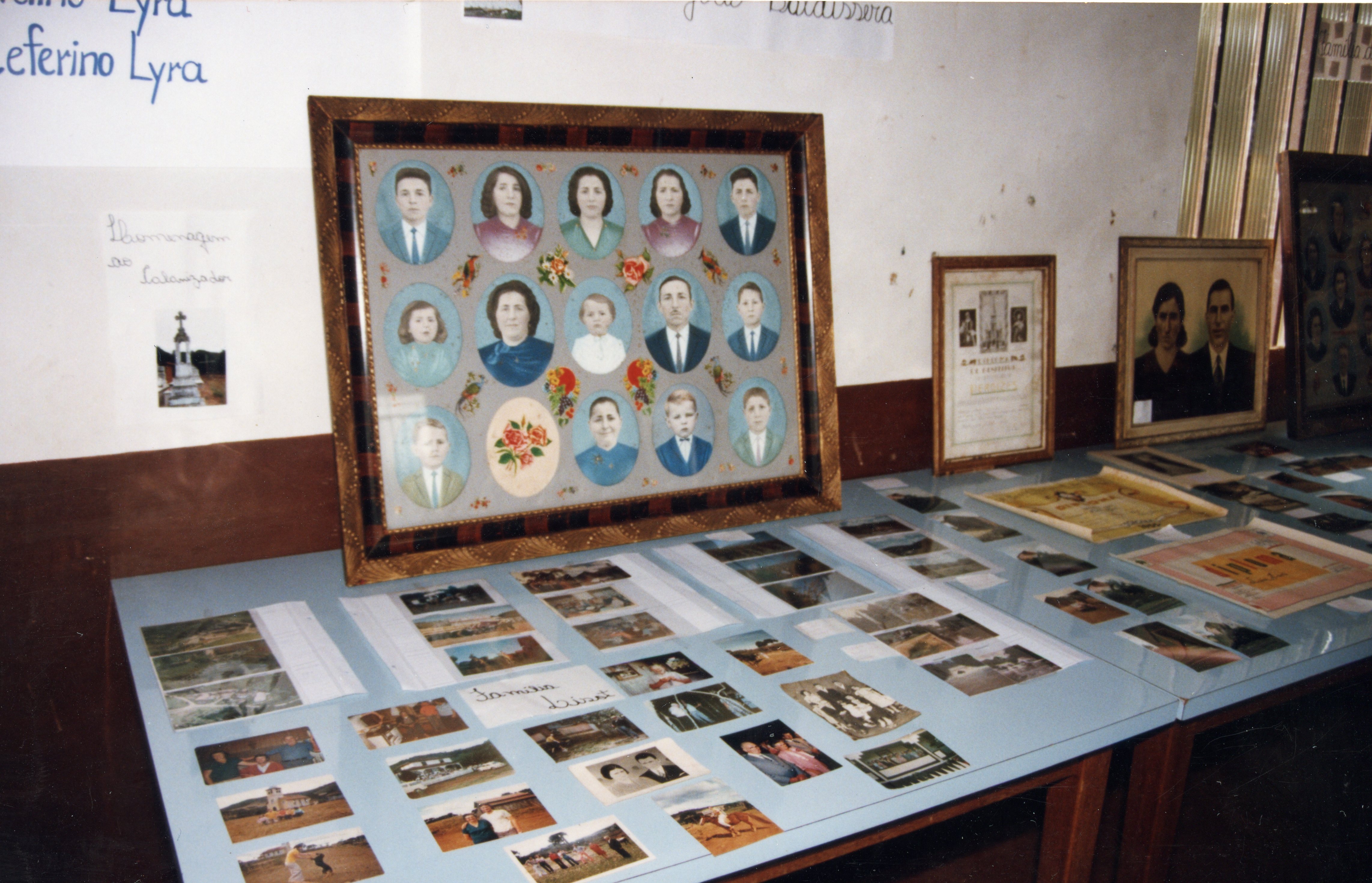 Uma exposição memorial ou arquivo histórico contendo um grande quadro com retratos de família em formato oval, acompanhado por diversas fotografias menores espalhadas sobre uma mesa. O ambiente sugere uma mostra cultural ou histórica, possivelmente em uma escola ou centro comunitário, preservando a memória de famílias locais.