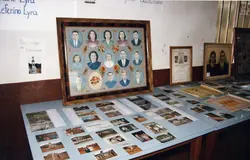 Uma exposição memorial ou arquivo histórico contendo um grande quadro com retratos de família em formato oval, acompanhado por diversas fotografias menores espalhadas sobre uma mesa. O ambiente sugere uma mostra cultural ou histórica, possivelmente em uma escola ou centro comunitário, preservando a memória de famílias locais.