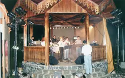 Uma banda de jovens se apresenta em um coreto de madeira durante um evento noturno. A cena, que remete aos anos 90, mostra músicos com instrumentos e equipamentos de som em uma estrutura rústica, possivelmente em uma praça pública de uma cidade do interior.