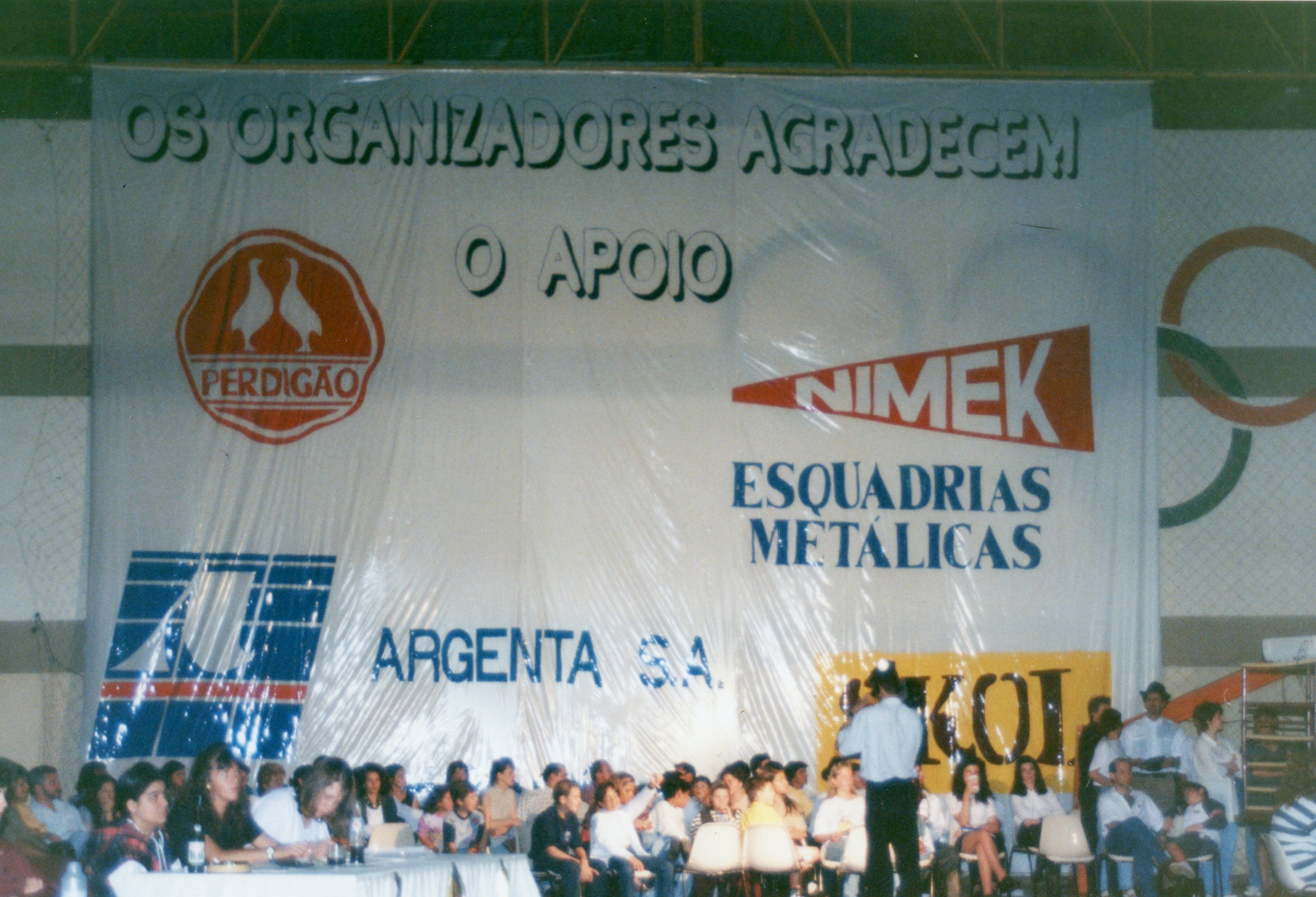 Uma faixa de grandes dimensões exibindo logotipos de empresas locais como Perdigão, Nimek e Argenta S.A., indicando um evento patrocinado ou uma cerimônia pública. Abaixo da faixa, observa-se um grupo de pessoas sentadas em um ambiente interno, possivelmente um ginásio, sugerindo um evento comunitário ou esportivo em uma cidade brasileira como Videira.