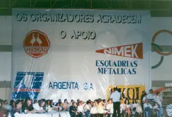 Uma faixa de grandes dimensões exibindo logotipos de empresas locais como Perdigão, Nimek e Argenta S.A., indicando um evento patrocinado ou uma cerimônia pública. Abaixo da faixa, observa-se um grupo de pessoas sentadas em um ambiente interno, possivelmente um ginásio, sugerindo um evento comunitário ou esportivo em uma cidade brasileira como Videira.
