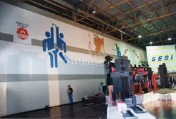 Uma fotografia interna de um ginásio de esportes decorado para um evento, possivelmente em Videira, dada a presença da marca Perdigão. O ambiente apresenta faixas com logotipos da Perdigão e do SESI, além de um palco montado com equipamentos de som e uma pessoa cantando, sugerindo uma celebração ou evento comunitário ocorrido provavelmente na década de 1990.
