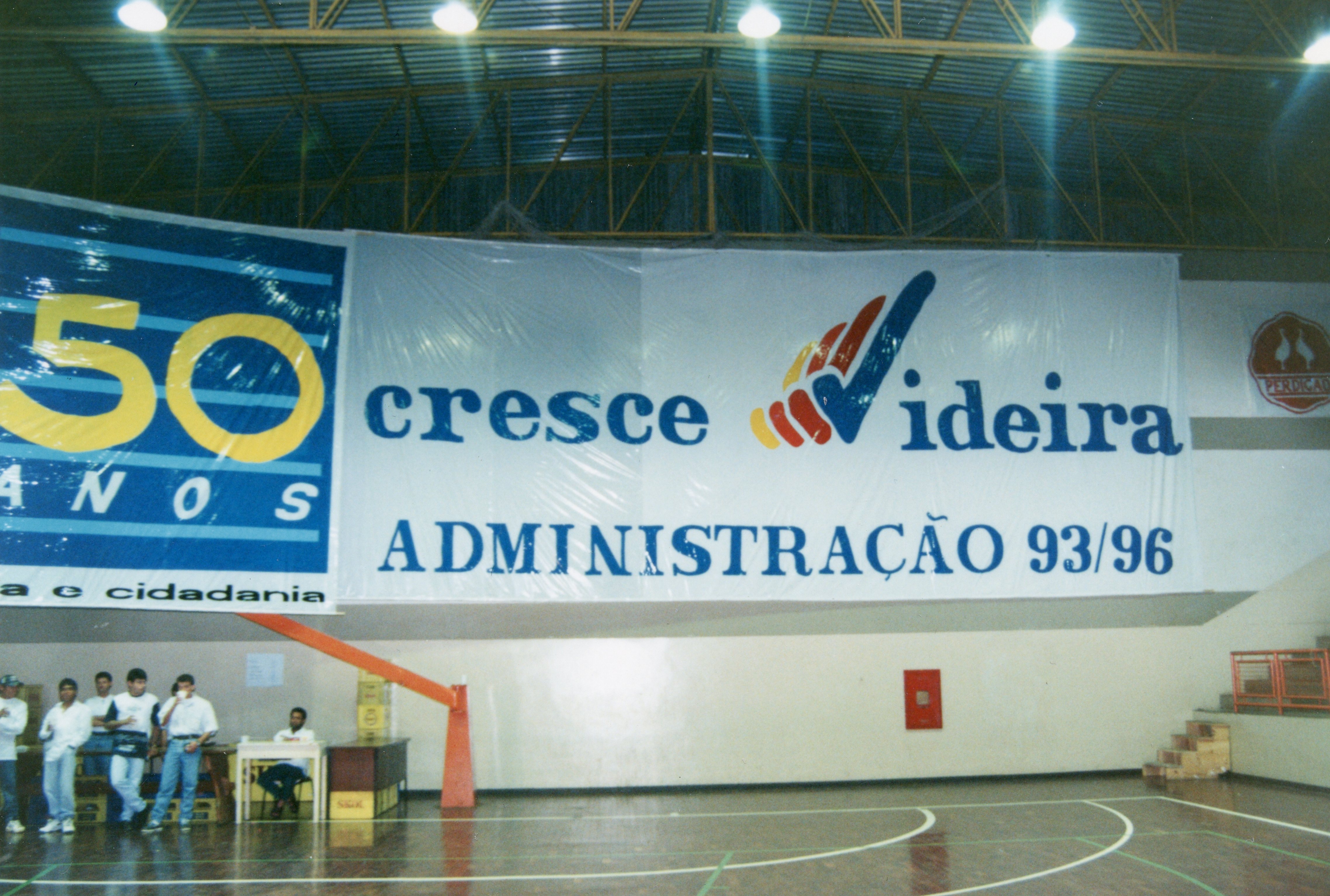 Uma fotografia interna de um ginásio de esportes em Videira, exibindo uma grande faixa comemorativa dos 50 anos do município e a gestão administrativa de 1993-1996. O ambiente apresenta uma estrutura esportiva típica com quadra poliesportiva e um grupo de pessoas reunidas próximo a uma mesa lateral.