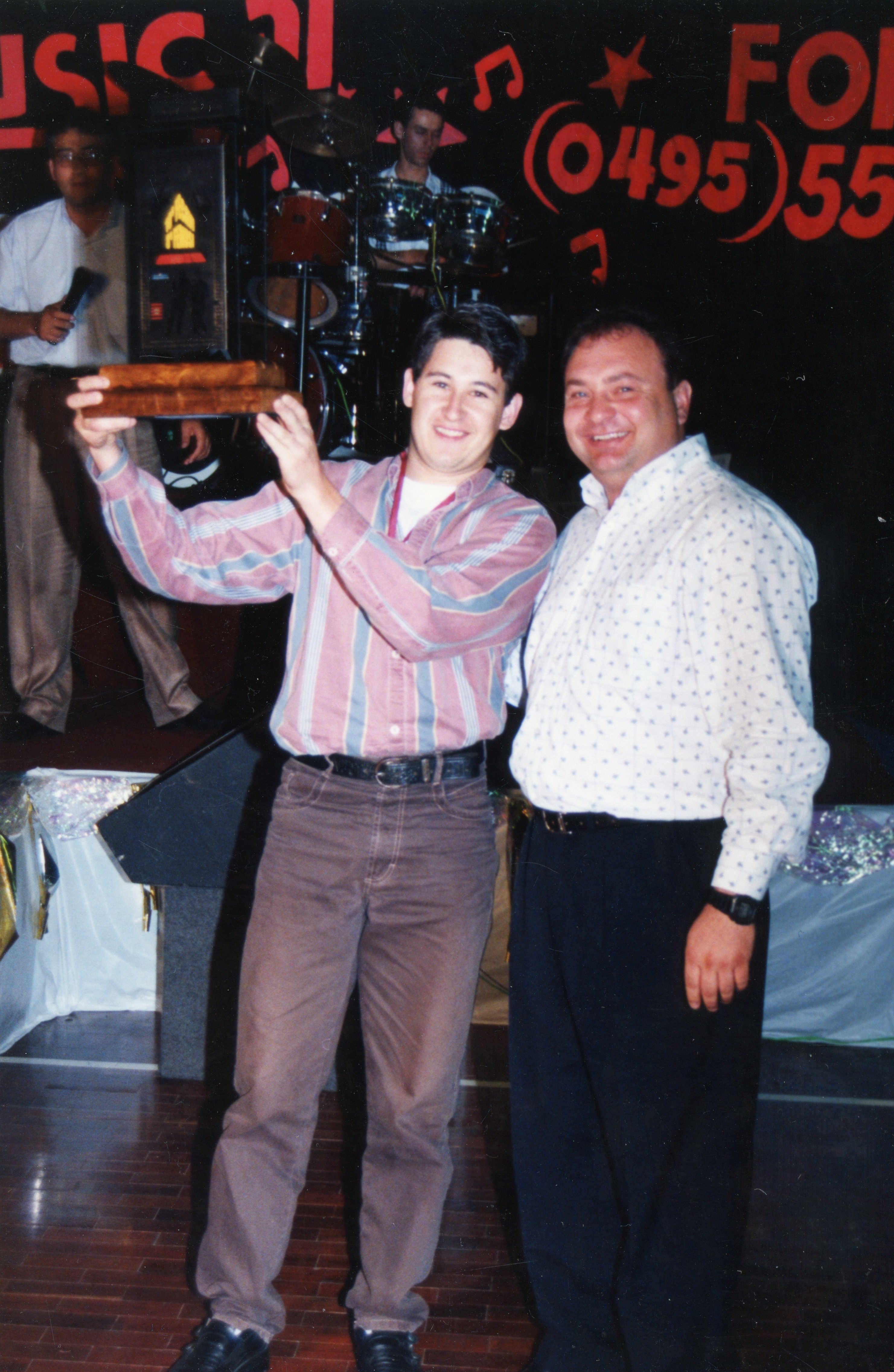 Fotografia de um evento social, possivelmente uma premiação musical, onde dois homens posam sorridentes segurando um troféu de madeira. Ao fundo, observa-se um palco com bateria e um banner com informações de contato, sugerindo um contexto de entretenimento ou celebração local, provavelmente na década de 1990.