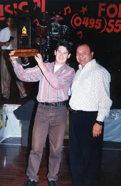 Fotografia de um evento social, possivelmente uma premiação musical, onde dois homens posam sorridentes segurando um troféu de madeira. Ao fundo, observa-se um palco com bateria e um banner com informações de contato, sugerindo um contexto de entretenimento ou celebração local, provavelmente na década de 1990.