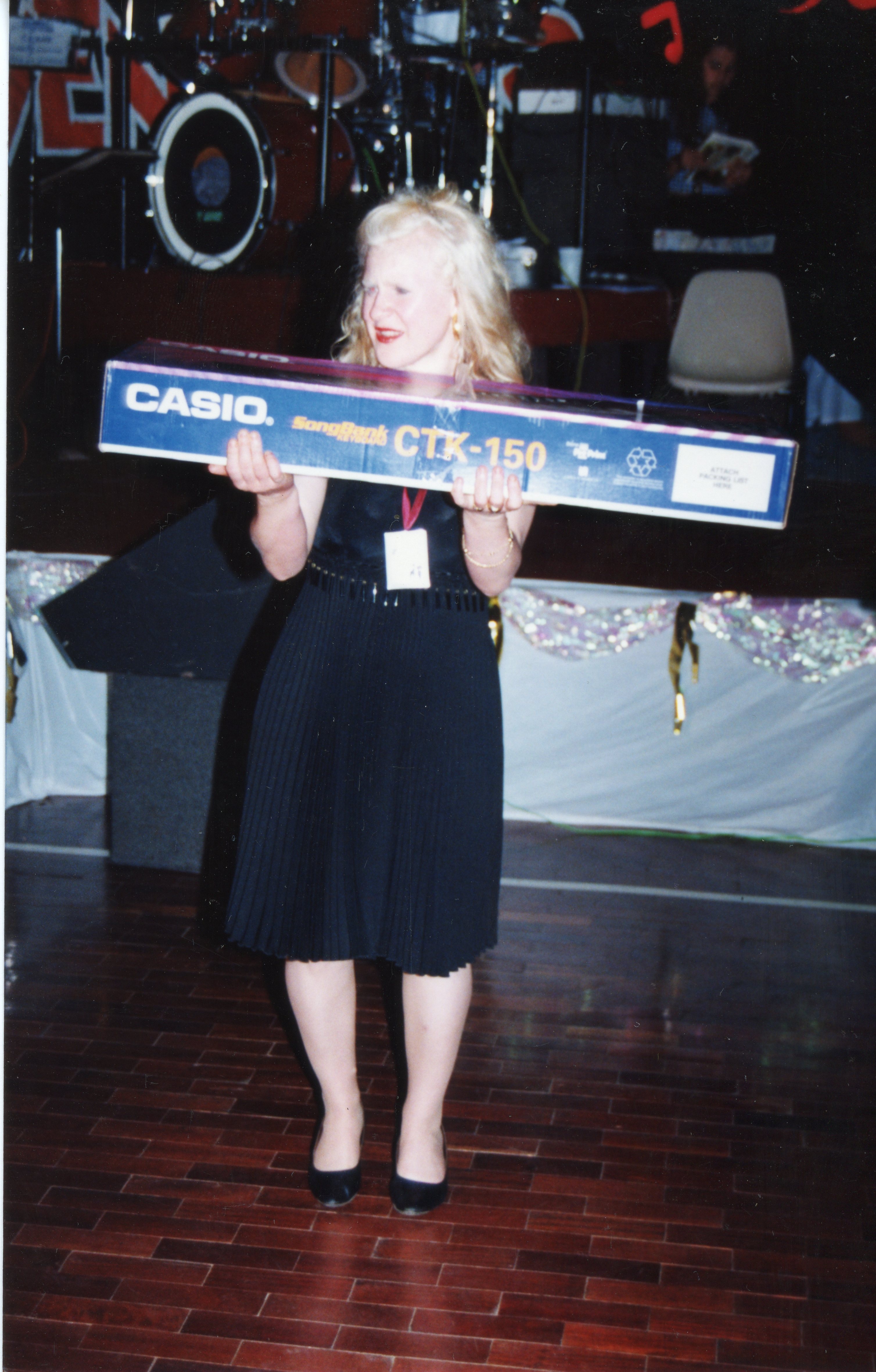 Uma mulher loira segura uma caixa de teclado Casio CTK-150 em um palco, possivelmente durante um evento de premiação ou sorteio. O cenário sugere um ambiente de salão de festas ou auditório, com instrumentos musicais ao fundo, típico de eventos sociais da década de 1990.