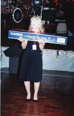 Uma mulher loira segura uma caixa de teclado Casio CTK-150 em um palco, possivelmente durante um evento de premiação ou sorteio. O cenário sugere um ambiente de salão de festas ou auditório, com instrumentos musicais ao fundo, típico de eventos sociais da década de 1990.