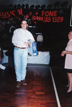 Uma fotografia de um evento social em ambiente interno, possivelmente um sorteio ou premiação, onde um homem segura uma caixa de teclado Casio. O cenário apresenta um palco com decoração festiva e um número de telefone visível ao fundo, sugerindo um contexto comercial ou de entretenimento típico da década de 1990.