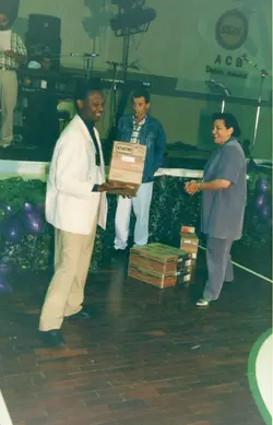 Uma fotografia colorida que registra um evento social ou promocional, possivelmente em um salão de festas, onde um homem entrega uma caixa de som da marca CCE para uma mulher. O ambiente apresenta decoração com balões e um banner da marca Skol ao fundo, sugerindo um contexto de premiação ou sorteio ocorrido provavelmente na década de 1990.