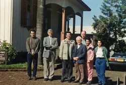 Uma fotografia colorida de um grupo de sete pessoas posando em frente a uma casa de madeira com arquitetura típica de colonização europeia, possivelmente em Videira. A vestimenta dos indivíduos sugere o final da década de 1980 ou início da década de 1990, com um carro modelo sedan estacionado ao fundo.
