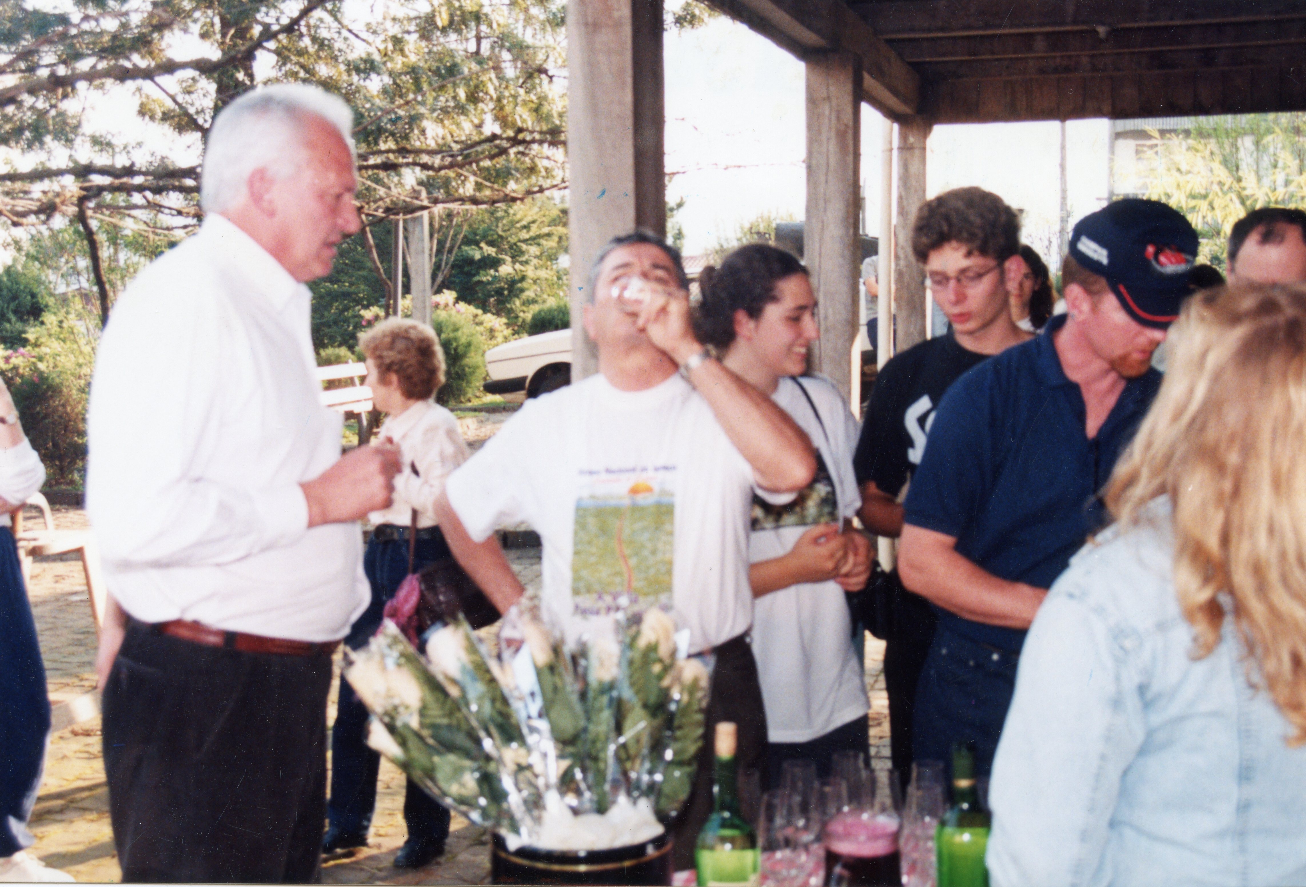 Uma cena de confraternização ao ar livre, possivelmente em um ambiente rural ou de lazer, com um grupo de pessoas reunidas em torno de uma mesa com bebidas e flores. A vestimenta e a qualidade da fotografia sugerem um registro casual do final da década de 1990 ou início dos anos 2000.