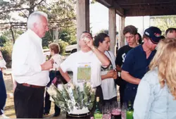 Uma cena de confraternização ao ar livre, possivelmente em um ambiente rural ou de lazer, com um grupo de pessoas reunidas em torno de uma mesa com bebidas e flores. A vestimenta e a qualidade da fotografia sugerem um registro casual do final da década de 1990 ou início dos anos 2000.