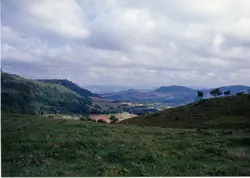 Uma paisagem rural com colinas verdes e um céu nublado. A vegetação é exuberante, com áreas de mata e campos cultivados em diferentes tons de verde e marrom. Ao fundo, montanhas se estendem até o horizonte. A imagem parece capturar um dia com nuvens densas, mas com luz solar filtrada, sugerindo um clima ameno. O cenário evoca uma sensação de tranquilidade e a vastidão da natureza.
