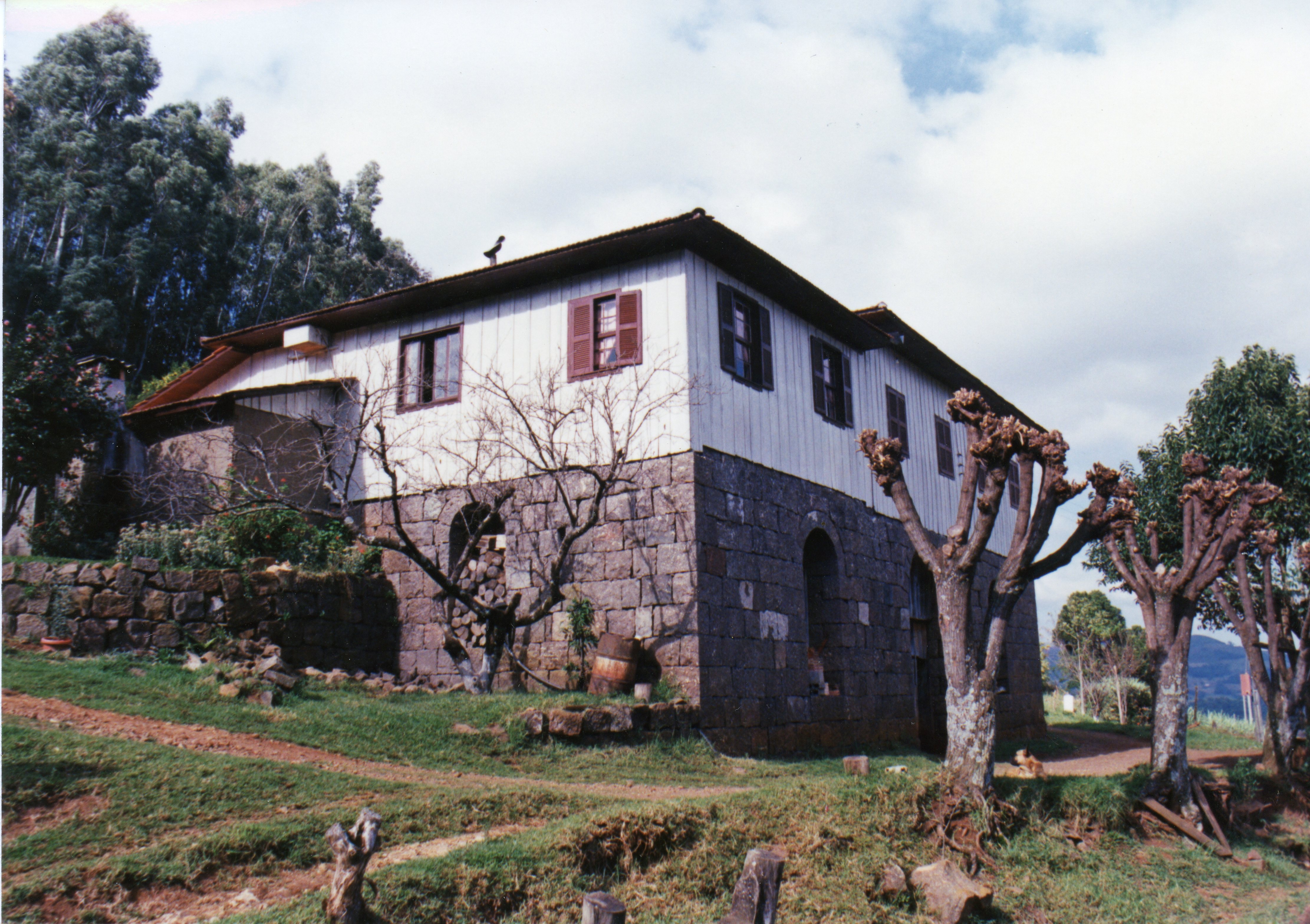 Uma casa rural tradicional com base de pedra e andar superior em madeira, típica da arquitetura de colonização europeia no sul do Brasil. A estrutura apresenta janelas com venezianas e um telhado de quatro águas, inserida em uma paisagem rural com árvores podadas e terreno inclinado.