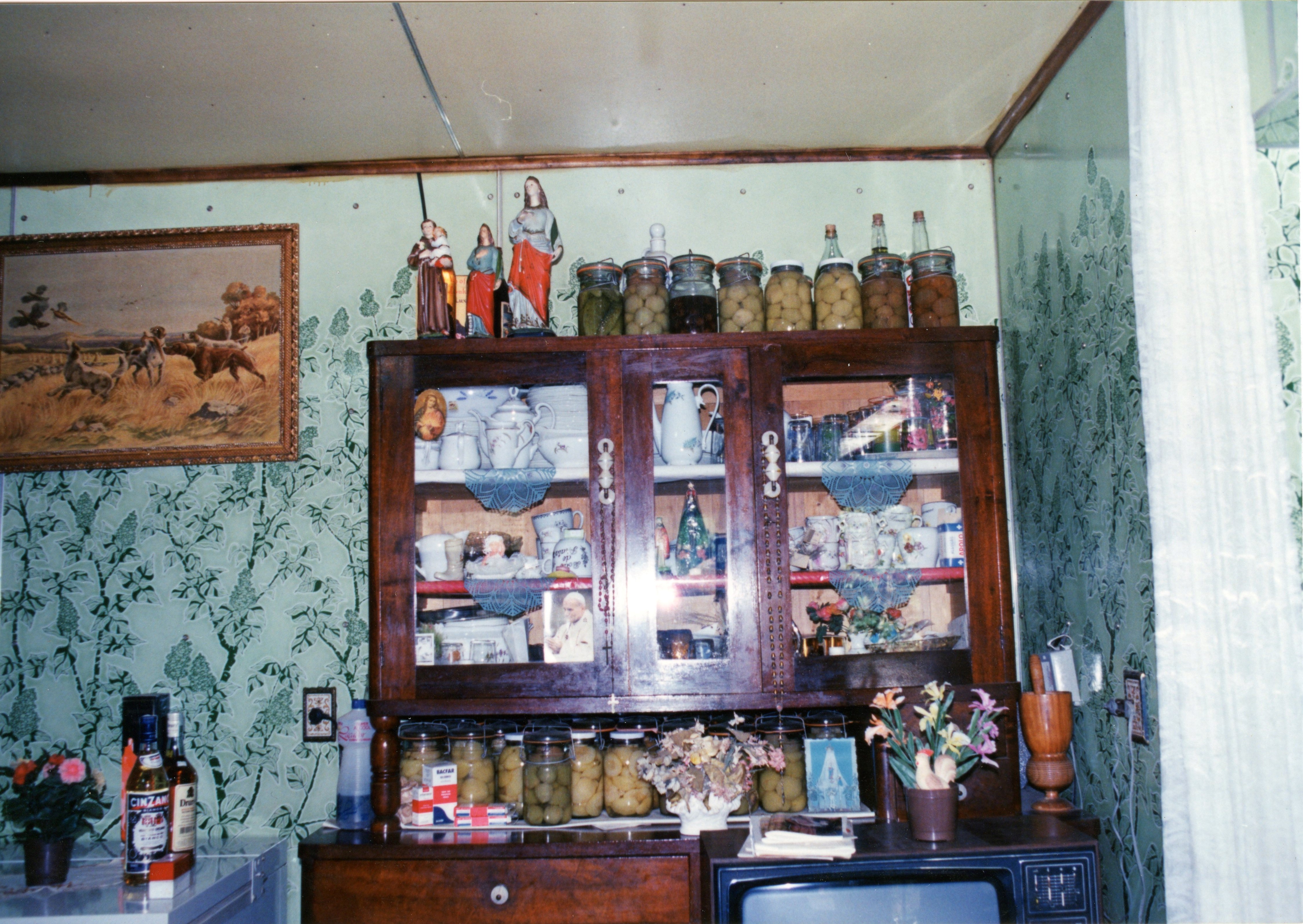 Uma fotografia de interior doméstico que exibe um armário de madeira rústico repleto de louças, conservas em compotas e objetos religiosos. O ambiente apresenta papel de parede com estampa floral e uma decoração típica de residências brasileiras de meados do século XX, sugerindo um contexto de vida rural ou de imigrantes.