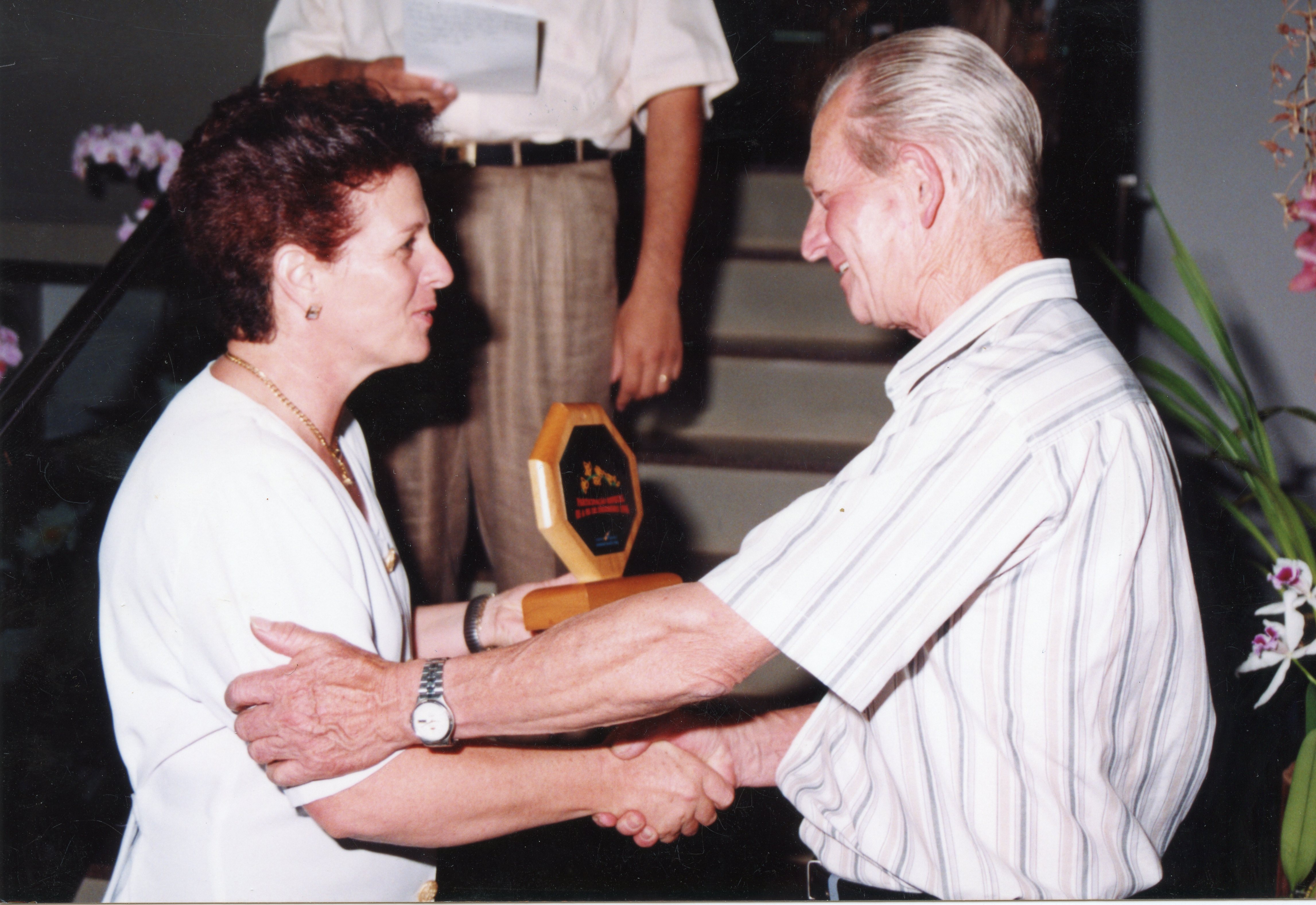Uma fotografia colorida que registra o momento de uma homenagem ou premiação, onde um homem entrega uma placa de madeira a uma mulher. O cenário sugere um ambiente interno, possivelmente um evento social ou corporativo, com vestimentas casuais típicas das décadas de 1990 ou 2000.