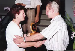Uma fotografia colorida que registra o momento de uma homenagem ou premiação, onde um homem entrega uma placa de madeira a uma mulher. O cenário sugere um ambiente interno, possivelmente um evento social ou corporativo, com vestimentas casuais típicas das décadas de 1990 ou 2000.