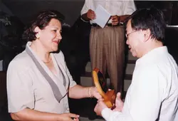 Uma fotografia que registra o momento de entrega de uma placa de homenagem ou premiação entre uma mulher e um homem. O cenário sugere um ambiente interno, possivelmente um evento corporativo ou social, com vestimentas formais típicas do final do século XX.