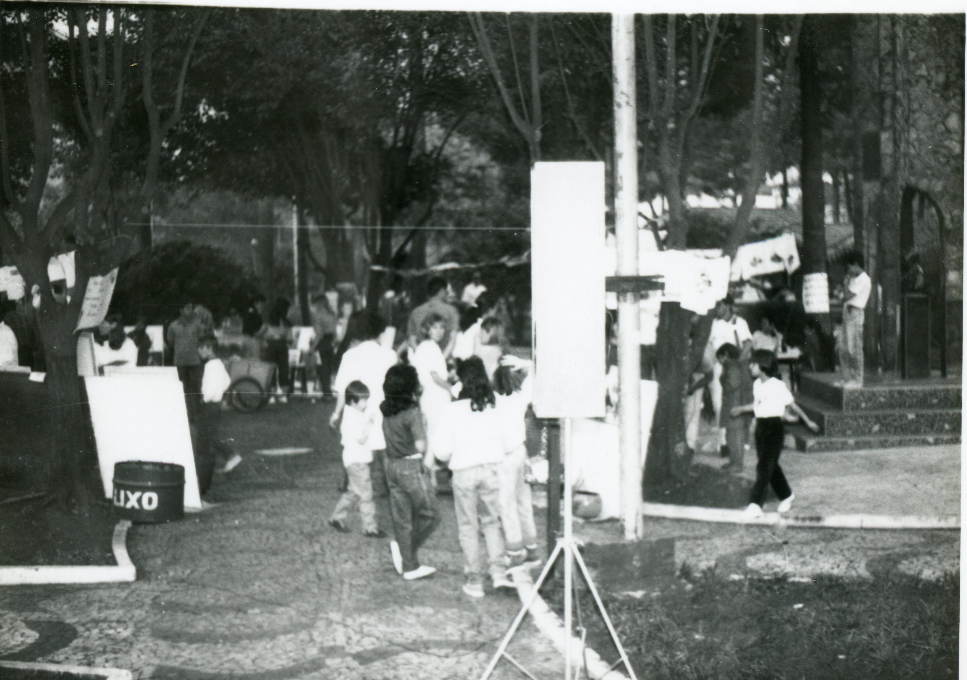 Fotografia em preto e branco de uma praça pública movimentada, possivelmente durante um evento comunitário ou feira cultural. Observam-se várias pessoas, incluindo crianças, circulando entre árvores e estruturas temporárias, com vestimentas que sugerem o final do século XX.