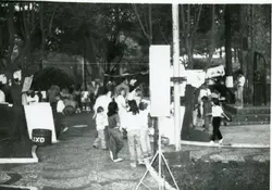 Fotografia em preto e branco de uma praça pública movimentada, possivelmente durante um evento comunitário ou feira cultural. Observam-se várias pessoas, incluindo crianças, circulando entre árvores e estruturas temporárias, com vestimentas que sugerem o final do século XX.