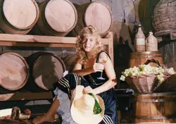 Uma jovem mulher loira posa em uma adega rústica, cercada por barris de madeira e cestas de uvas, segurando uma taça de vinho. A cena evoca a cultura da vitivinicultura, possivelmente em uma região como Videira, com a mulher vestindo trajes que remetem a festividades tradicionais ou folclóricas.