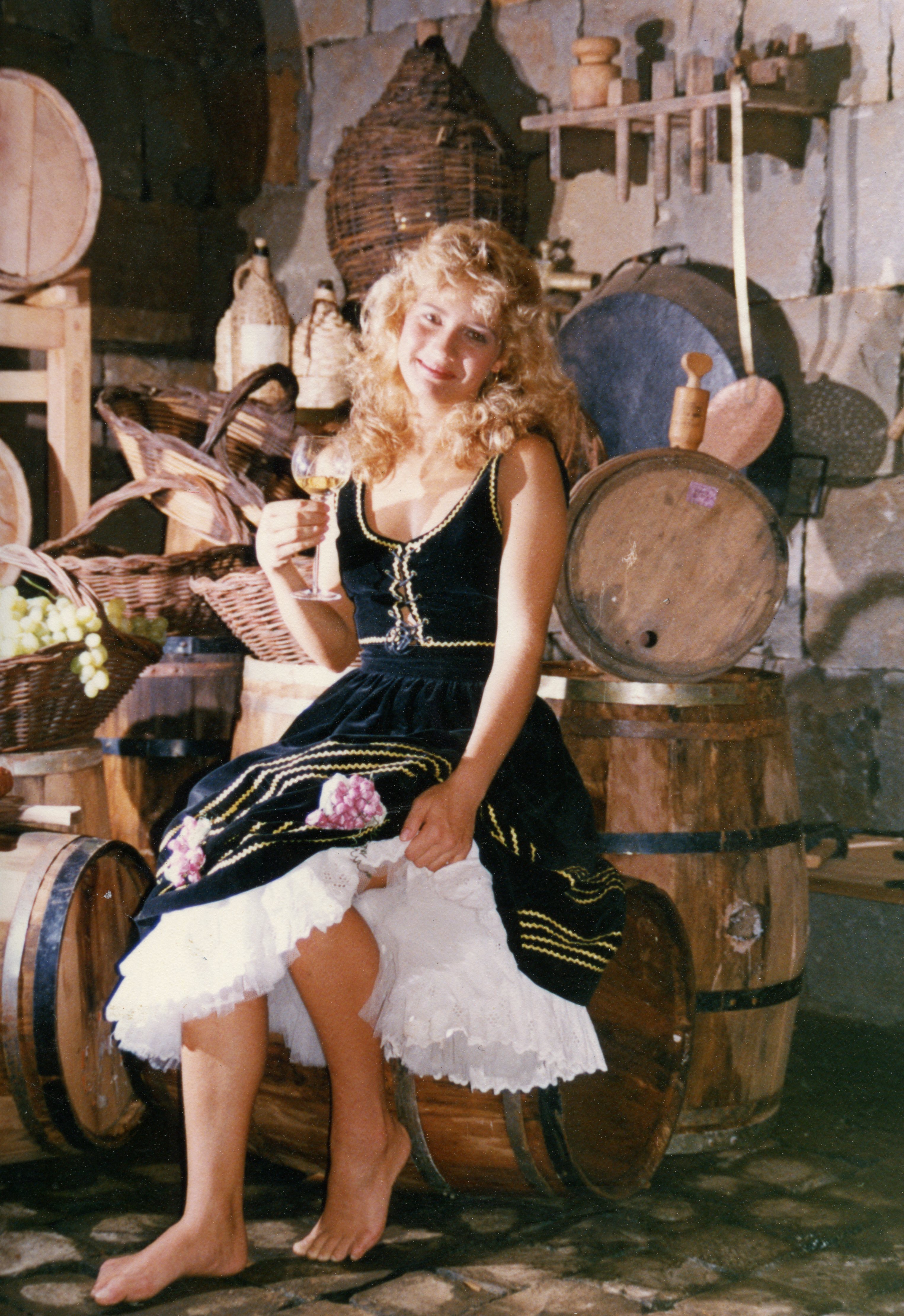 Uma jovem loira vestida com trajes típicos de inspiração germânica, segurando uma taça de vinho, sentada sobre um barril de madeira em uma adega rústica. O cenário evoca a cultura da vitivinicultura, possivelmente associada a festas tradicionais ou eventos de promoção de vinhos em regiões de colonização europeia no Brasil.
