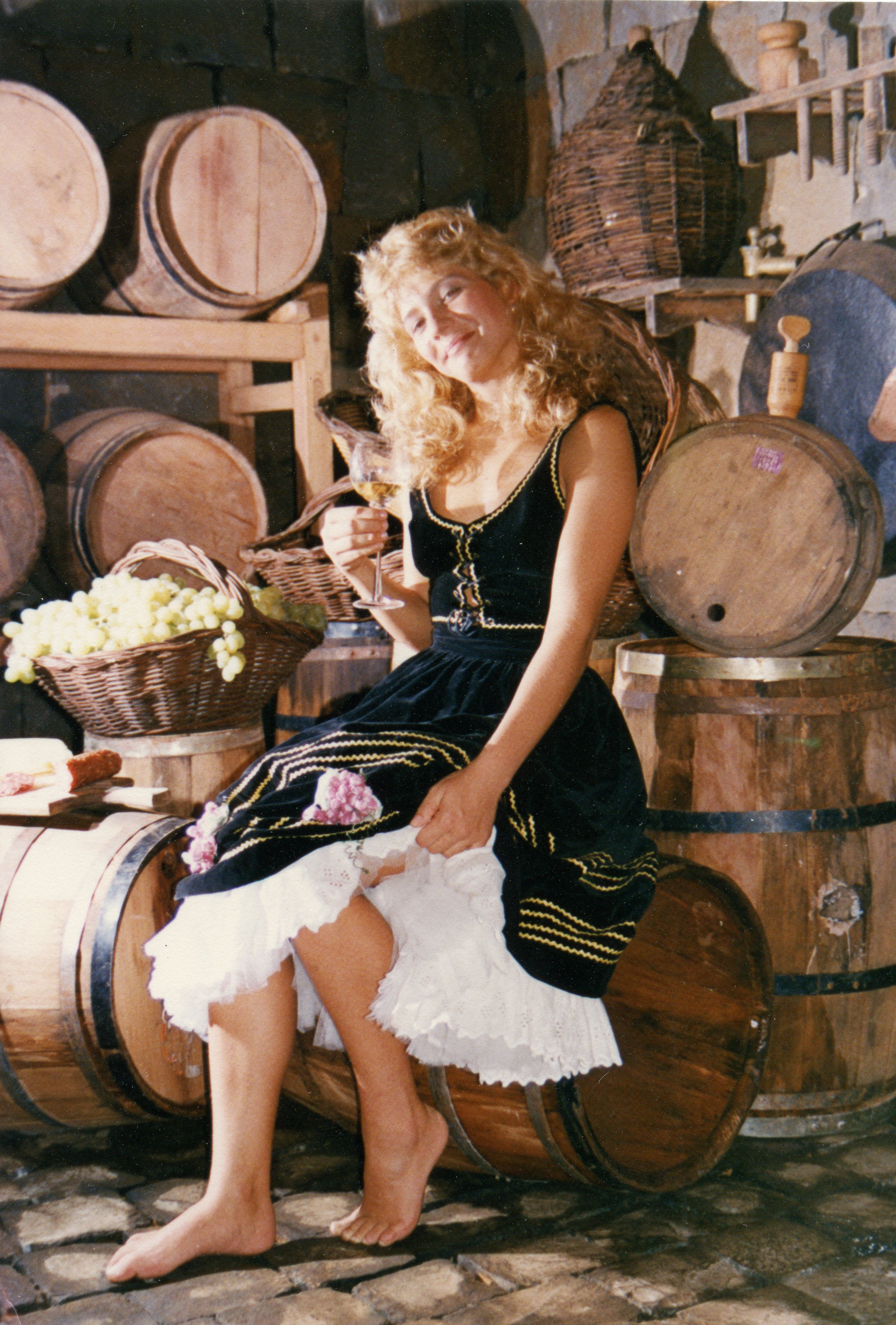 Uma mulher jovem com cabelos loiros veste um traje típico de inspiração germânica, sentada sobre um barril de madeira em uma adega rústica. O cenário é composto por diversos barris de vinho e cestas de uvas, sugerindo uma celebração cultural ou promoção turística relacionada à vitivinicultura, possivelmente em uma região de colonização europeia como Videira.