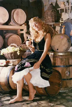 Uma mulher jovem com cabelos loiros veste um traje típico de inspiração germânica, sentada sobre um barril de madeira em uma adega rústica. O cenário é composto por diversos barris de vinho e cestas de uvas, sugerindo uma celebração cultural ou promoção turística relacionada à vitivinicultura, possivelmente em uma região de colonização europeia como Videira.
