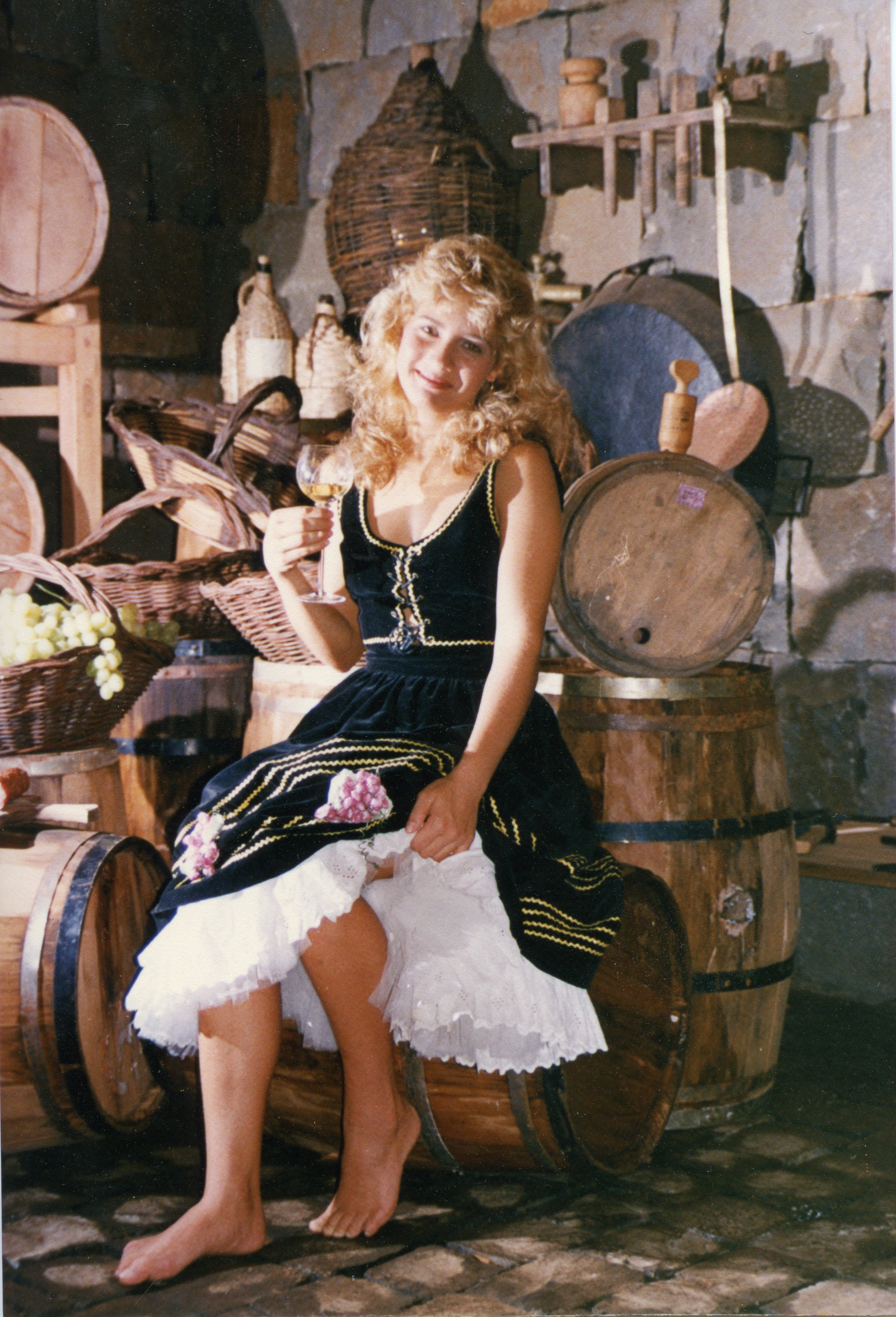 Uma jovem loira vestida com trajes típicos de inspiração tirolesa ou germânica segura uma taça de vinho, sentada sobre um barril de madeira em uma adega rústica. O cenário, repleto de cestos de vime, uvas e barris, evoca a cultura da vitivinicultura, possivelmente associada a festividades como a Festa da Uva em regiões de colonização europeia no Brasil.