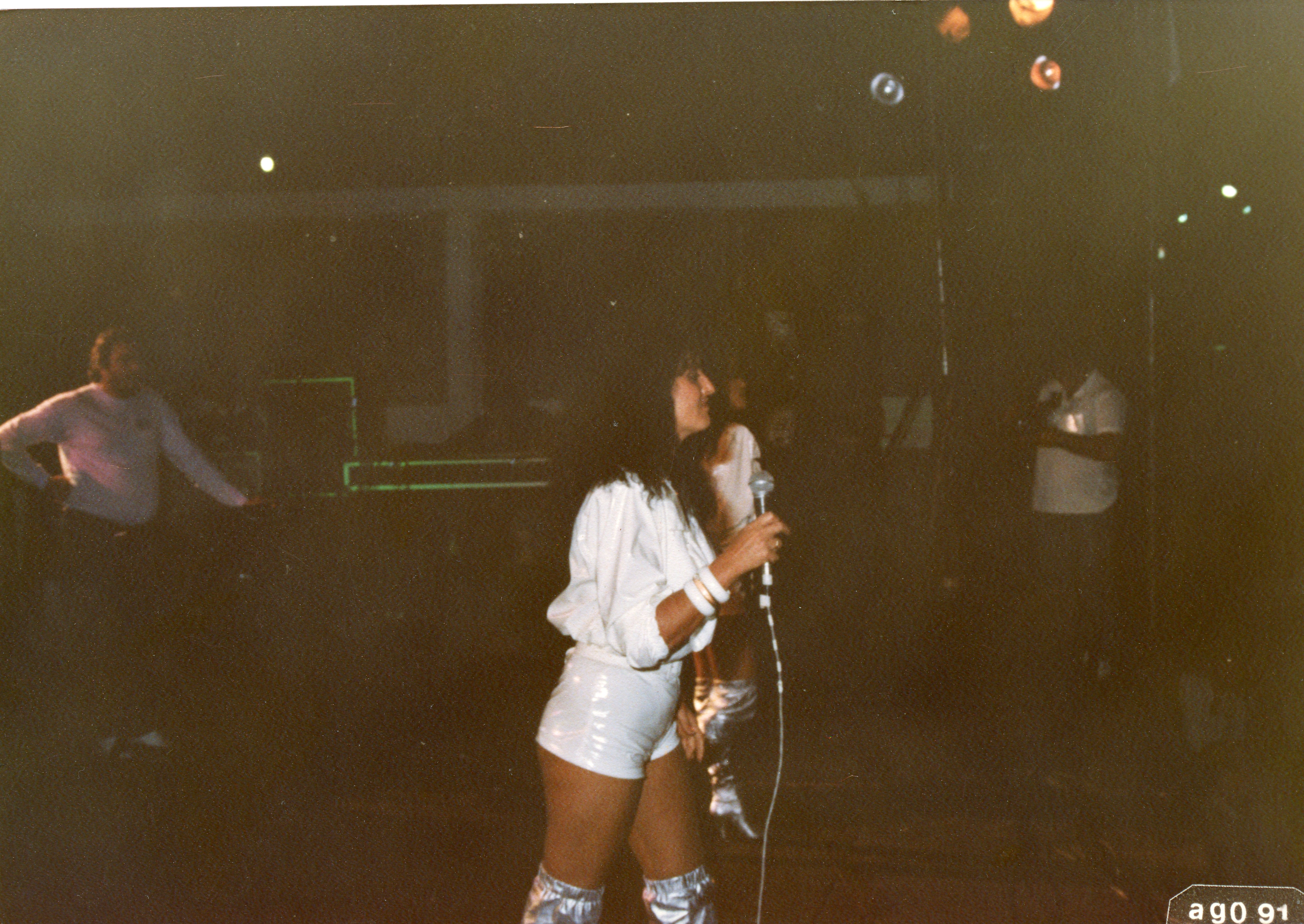 Uma fotografia de uma apresentação musical em um ambiente noturno, possivelmente um clube ou salão de festas. A cantora está em destaque, vestindo roupas brancas e botas prateadas, segurando um microfone, com outras pessoas ao fundo em um cenário de baixa luminosidade.