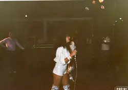 Uma fotografia de uma apresentação musical em um ambiente noturno, possivelmente um clube ou salão de festas. A cantora está em destaque, vestindo roupas brancas e botas prateadas, segurando um microfone, com outras pessoas ao fundo em um cenário de baixa luminosidade.