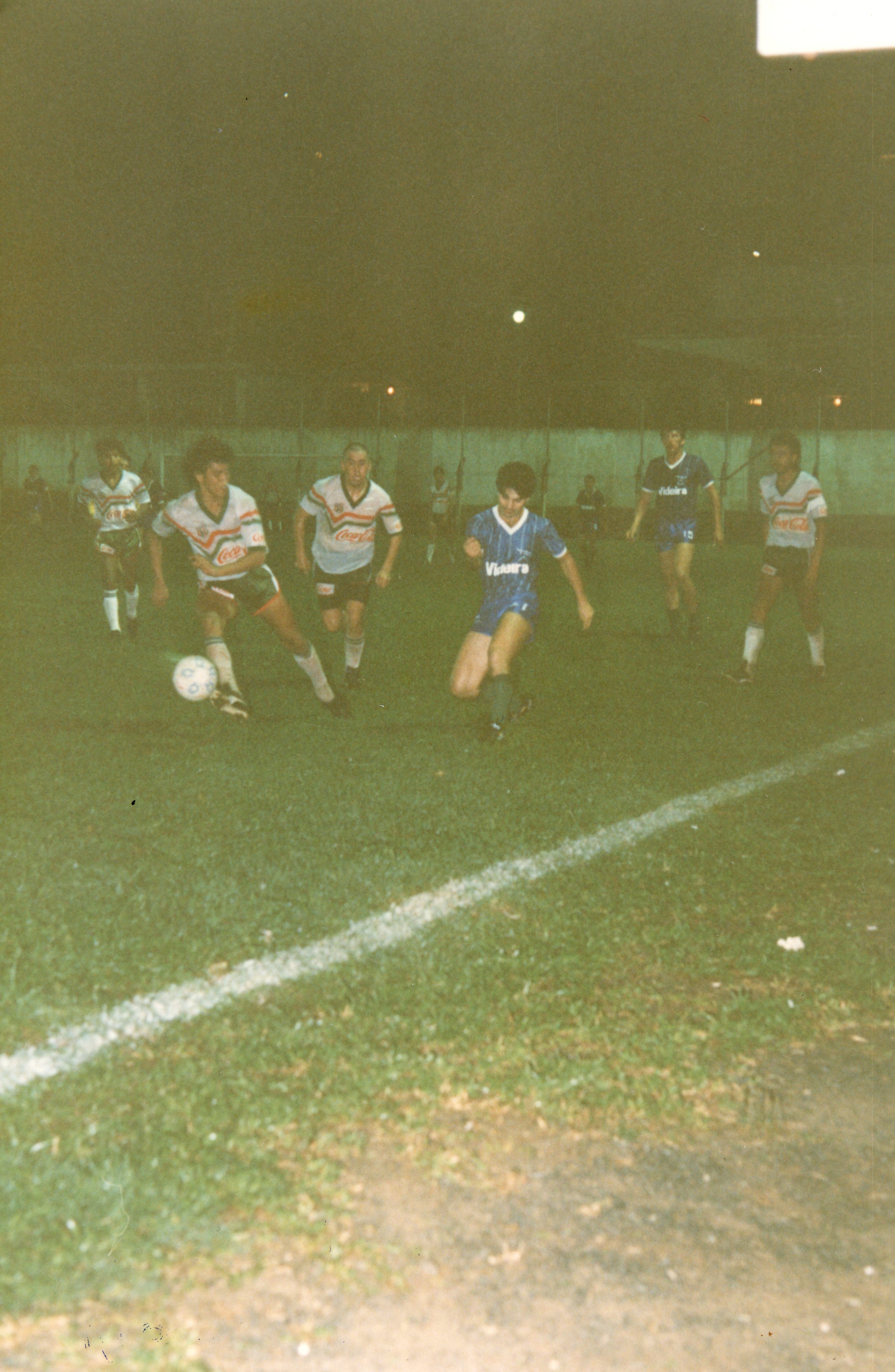 Uma partida de futebol amador noturna em um campo de grama, possivelmente em Videira, Santa Catarina. Os jogadores vestem uniformes típicos da década de 1980 ou 1990, com patrocínios visíveis, em uma cena de jogo dinâmico sob iluminação artificial.