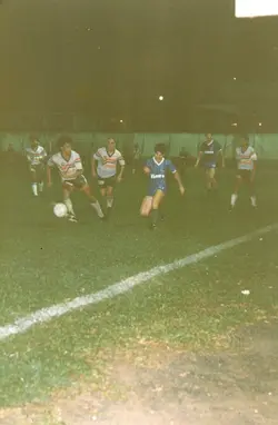 Uma partida de futebol amador noturna em um campo de grama, possivelmente em Videira, Santa Catarina. Os jogadores vestem uniformes típicos da década de 1980 ou 1990, com patrocínios visíveis, em uma cena de jogo dinâmico sob iluminação artificial.