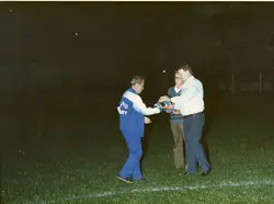 Uma fotografia noturna capturando uma cerimônia de entrega de troféu ou homenagem em um campo de futebol. Três homens estão presentes, sendo que um deles veste um agasalho esportivo com a inscrição 'Grêmio', sugerindo um contexto de evento esportivo amador ou regional.