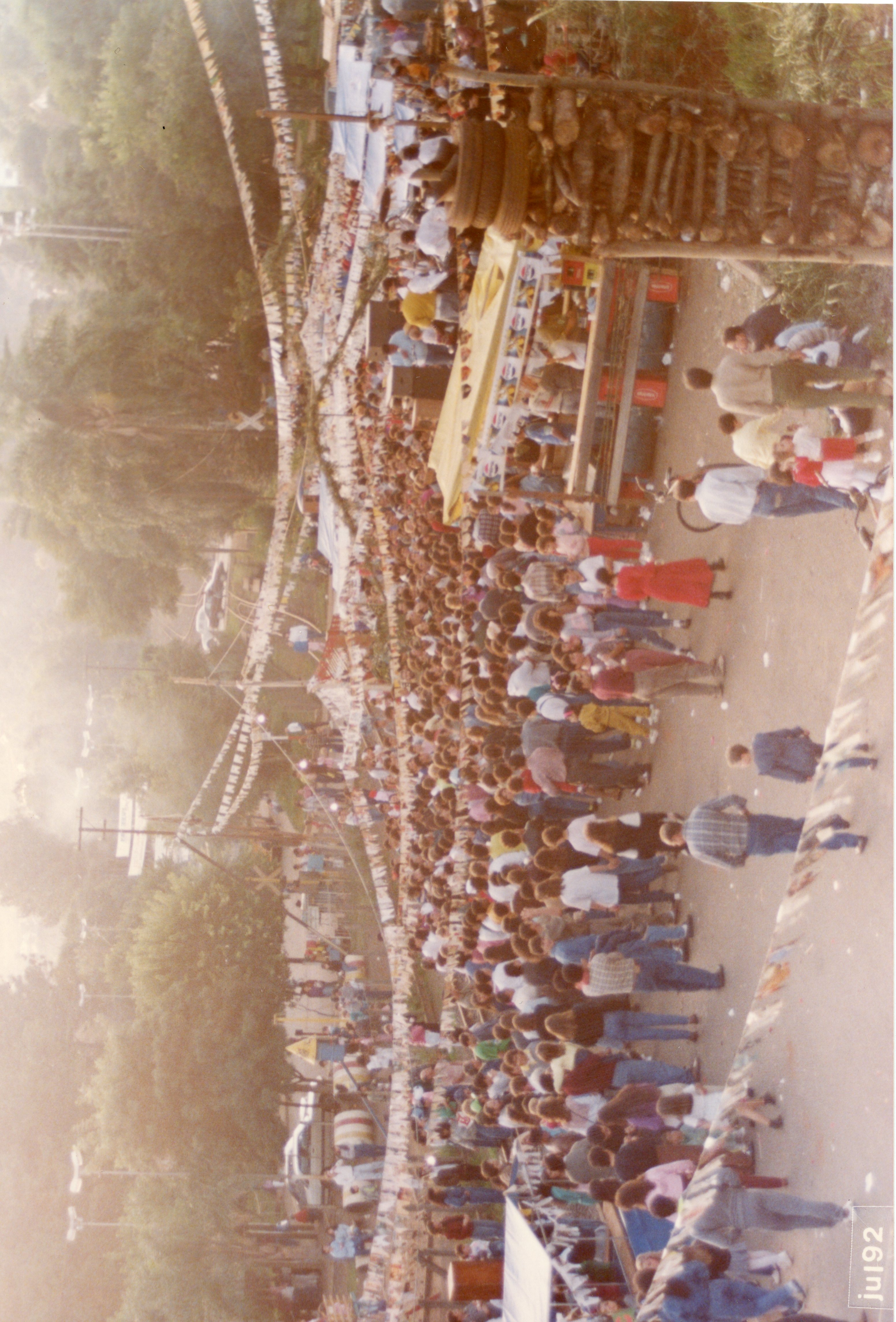 Uma fotografia aérea capturando uma grande aglomeração de pessoas em um evento ao ar livre, possivelmente uma festa popular ou quermesse, decorada com bandeirinhas típicas. A cena exibe barracas, uma estrutura de madeira empilhada e uma multidão vestindo roupas casuais típicas do início da década de 1990, sugerindo um contexto de celebração comunitária em uma cidade brasileira.