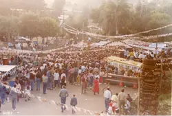 Uma fotografia aérea capturando uma grande aglomeração de pessoas em um evento ao ar livre, possivelmente uma festa popular ou quermesse, decorada com bandeirinhas típicas. A cena exibe barracas, uma estrutura de madeira empilhada e uma multidão vestindo roupas casuais típicas do início da década de 1990, sugerindo um contexto de celebração comunitária em uma cidade brasileira.