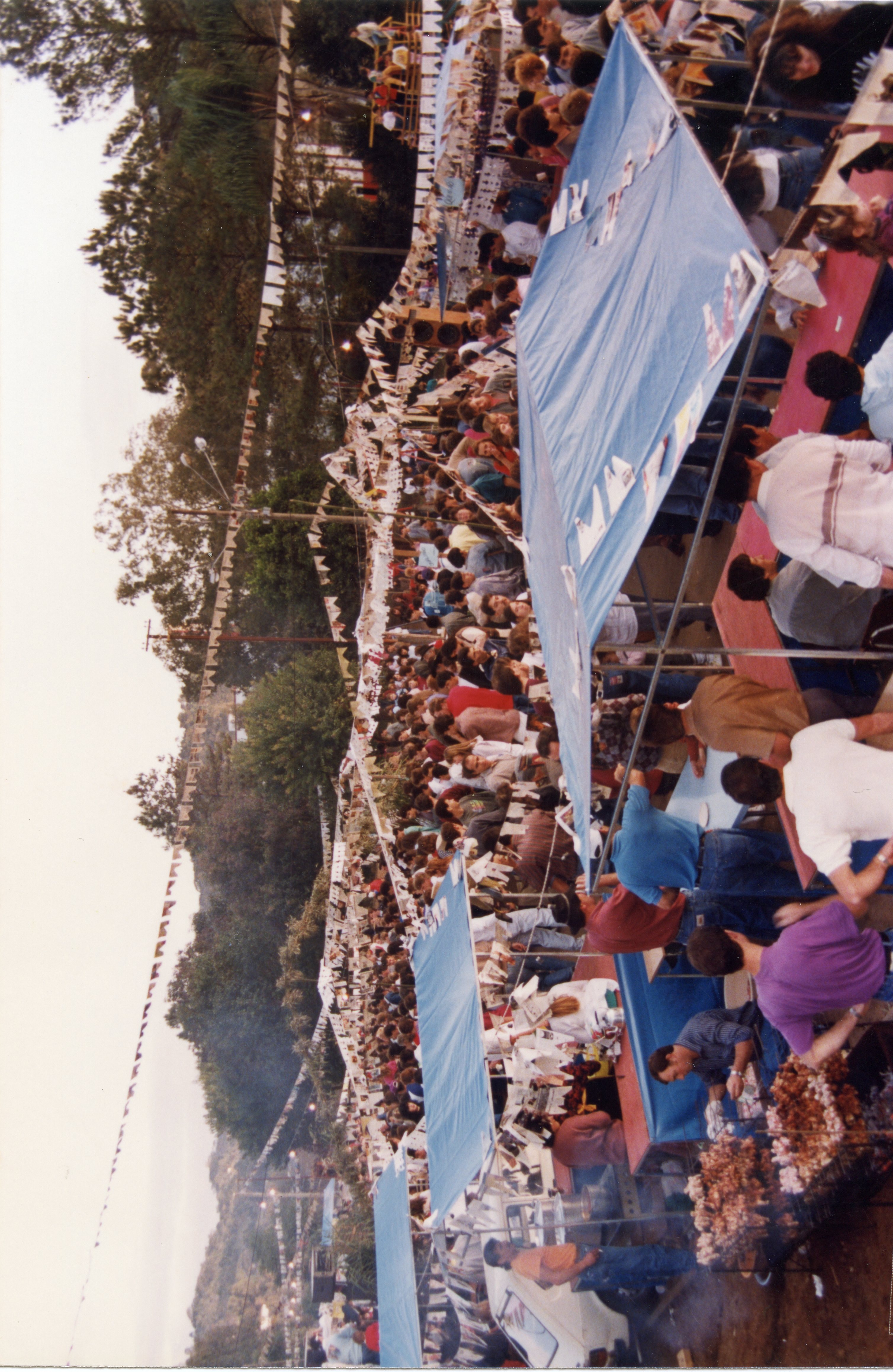 Uma fotografia de ângulo elevado capturando uma celebração popular ao ar livre, possivelmente uma festa junina ou evento comunitário, com diversas barracas cobertas por lonas azuis. O ambiente é decorado com bandeirolas típicas e apresenta uma grande aglomeração de pessoas, sugerindo um evento social tradicional em uma cidade brasileira.
