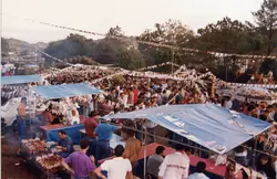 Uma fotografia de ângulo elevado capturando uma celebração popular ao ar livre, possivelmente uma festa junina ou evento comunitário, com diversas barracas cobertas por lonas azuis. O ambiente é decorado com bandeirolas típicas e apresenta uma grande aglomeração de pessoas, sugerindo um evento social tradicional em uma cidade brasileira.