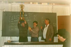 Uma fotografia colorida que registra a entrega de um troféu a um homem, em um ambiente interno com paredes de madeira. Ao fundo, um quadro-negro exibe anotações manuscritas sobre uma competição, possivelmente esportiva ou de rodeio, com nomes e pontuações, sugerindo um evento comunitário ou rural típico de pequenas cidades brasileiras.