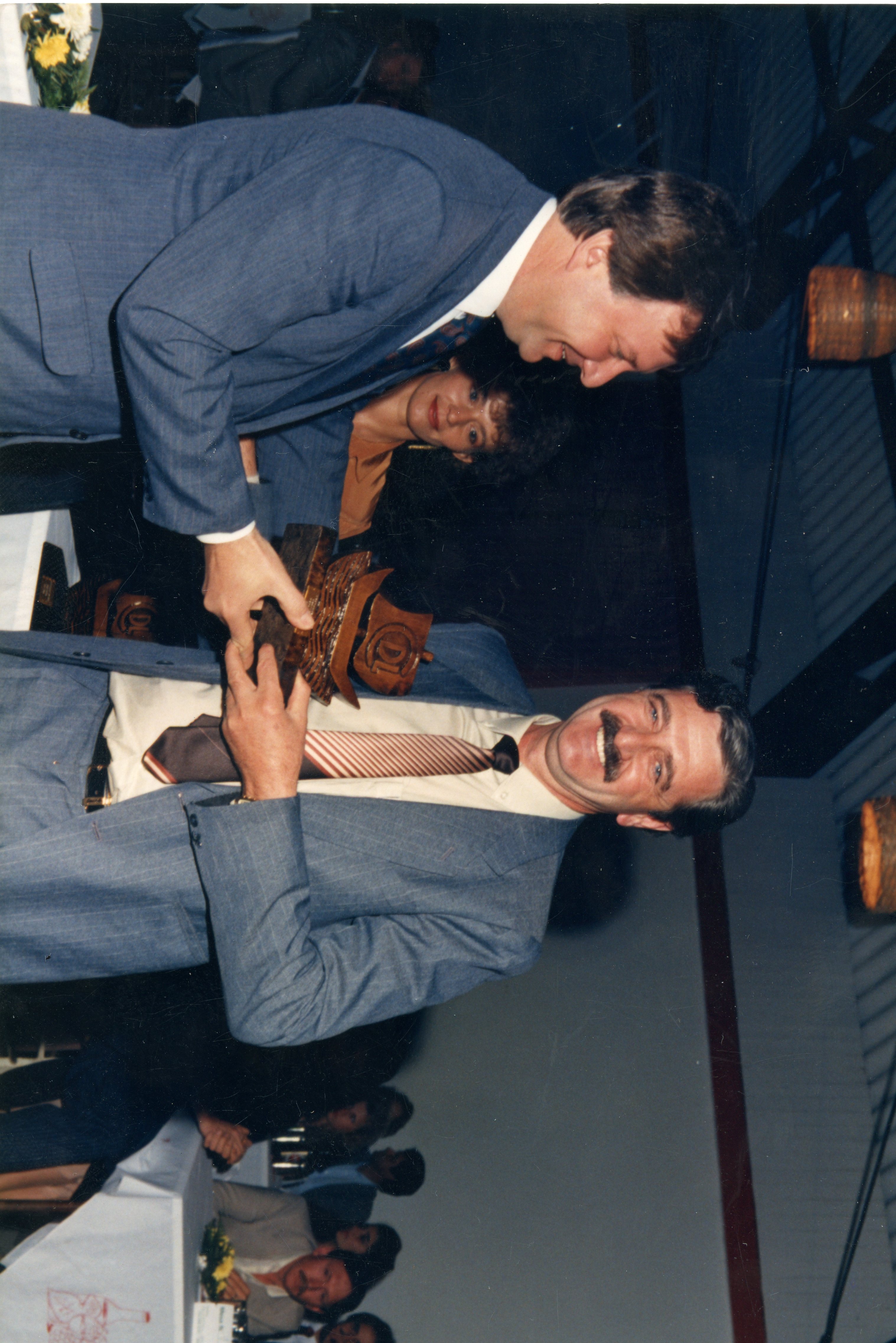 Uma fotografia colorida que registra um momento de premiação ou homenagem em um ambiente interno, possivelmente um evento social ou corporativo. Dois homens de terno e gravata estão em destaque, com um deles entregando um troféu de madeira, enquanto uma mulher observa ao fundo, sugerindo uma cerimônia formal típica das décadas de 1980 ou 1990.
