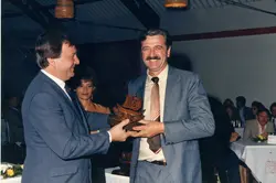 Uma fotografia colorida que registra um momento de premiação ou homenagem em um ambiente interno, possivelmente um evento social ou corporativo. Dois homens de terno e gravata estão em destaque, com um deles entregando um troféu de madeira, enquanto uma mulher observa ao fundo, sugerindo uma cerimônia formal típica das décadas de 1980 ou 1990.