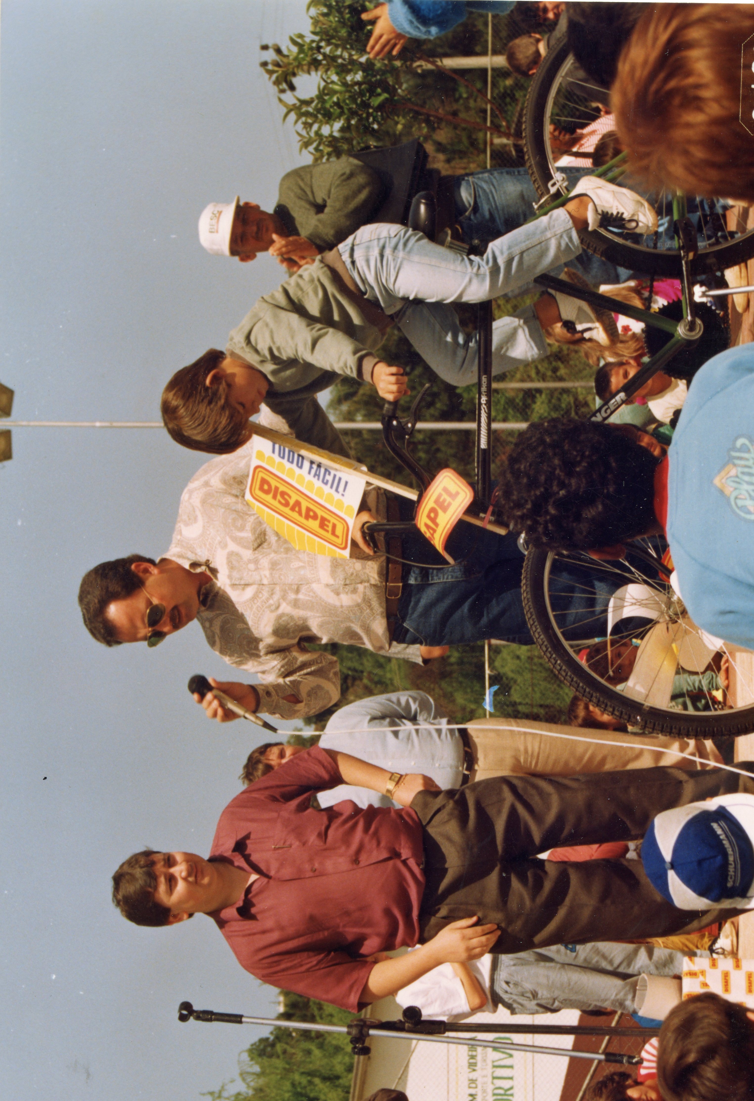 Uma fotografia colorida que registra um evento público ao ar livre, possivelmente uma competição ou gincana esportiva, com a presença de um locutor segurando um microfone. O cenário inclui bicicletas e pessoas vestindo roupas típicas da década de 1990, sugerindo um evento comunitário em uma cidade brasileira como Videira.