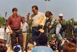 Uma fotografia colorida que registra um evento público ao ar livre, possivelmente uma competição ou gincana esportiva, com a presença de um locutor segurando um microfone. O cenário inclui bicicletas e pessoas vestindo roupas típicas da década de 1990, sugerindo um evento comunitário em uma cidade brasileira como Videira.