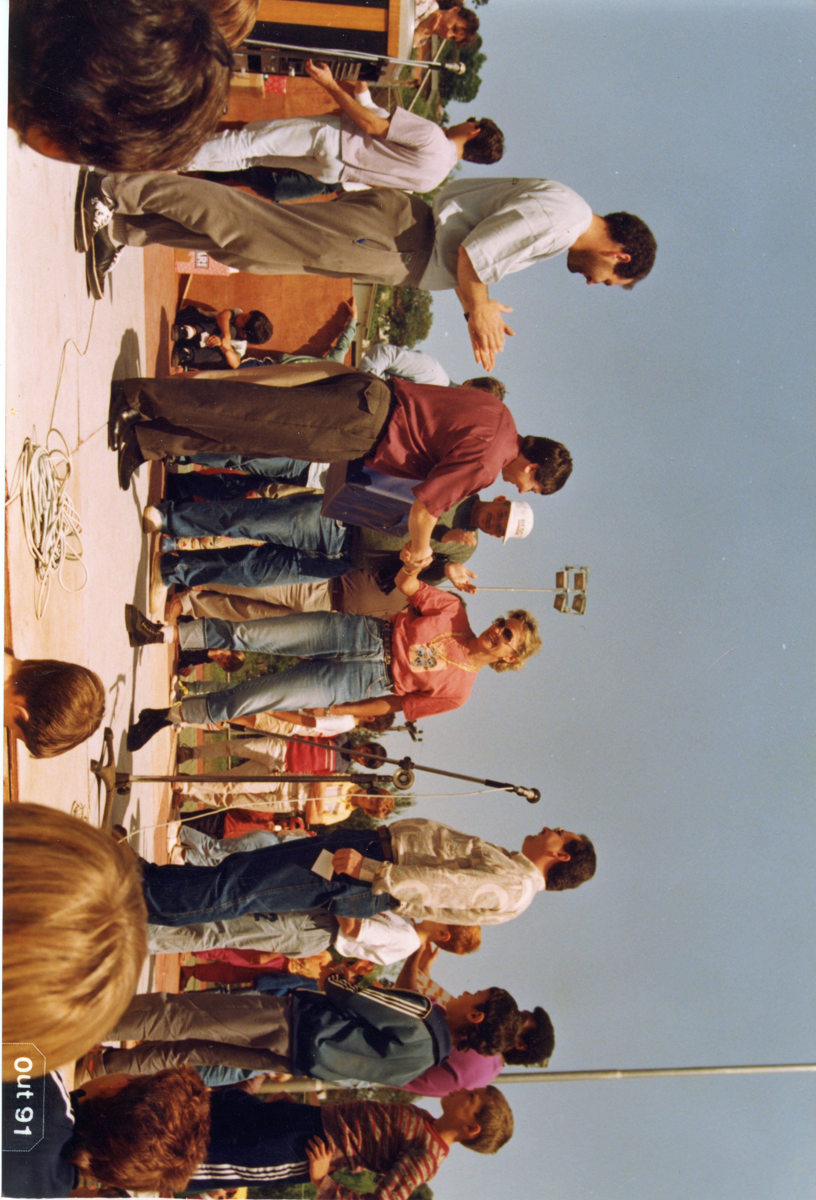 Fotografia colorida que registra um evento público ao ar livre, possivelmente uma cerimônia de premiação ou entrega de honrarias. As pessoas vestem roupas casuais típicas do início da década de 1990, com presença de microfones e equipamentos de som, sugerindo uma celebração comunitária.