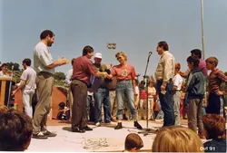 Fotografia colorida que registra um evento público ao ar livre, possivelmente uma cerimônia de premiação ou entrega de honrarias. As pessoas vestem roupas casuais típicas do início da década de 1990, com presença de microfones e equipamentos de som, sugerindo uma celebração comunitária.