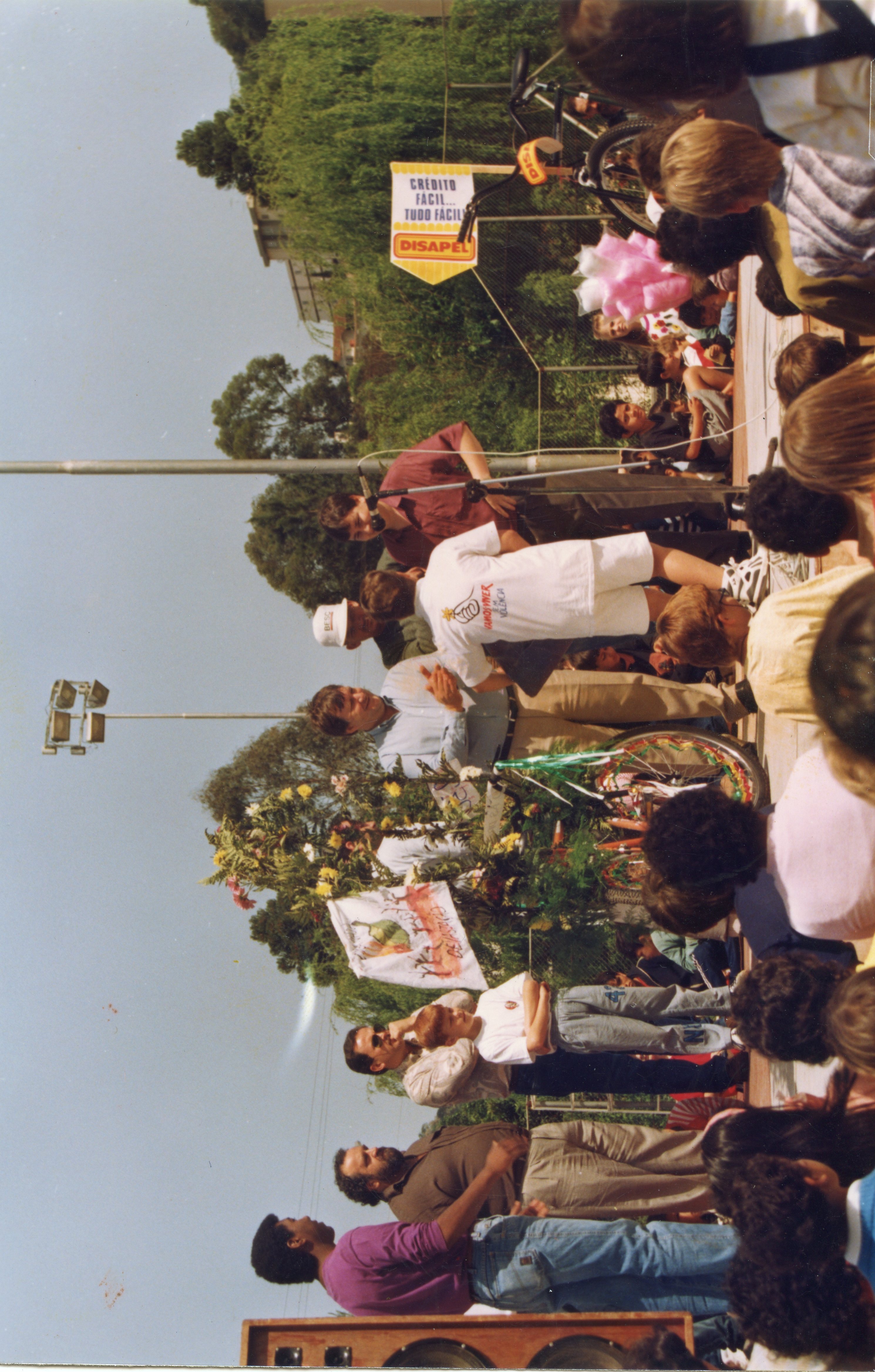 Uma fotografia colorida que registra um evento público ao ar livre, possivelmente uma celebração comunitária ou escolar em uma cidade brasileira. Observam-se pessoas de diversas idades, incluindo crianças e adultos, reunidas em torno de um palco ou área de apresentações, com elementos decorativos e uma placa publicitária da empresa 'Disapel'.