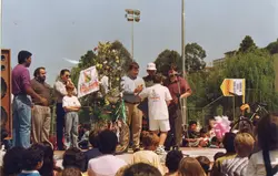 Uma fotografia colorida que registra um evento público ao ar livre, possivelmente uma celebração comunitária ou escolar em uma cidade brasileira. Observam-se pessoas de diversas idades, incluindo crianças e adultos, reunidas em torno de um palco ou área de apresentações, com elementos decorativos e uma placa publicitária da empresa 'Disapel'.